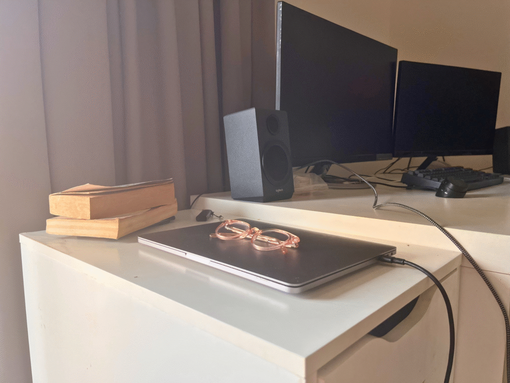 A laptop and books on a chest of drawers, next to a desk with two monitors.